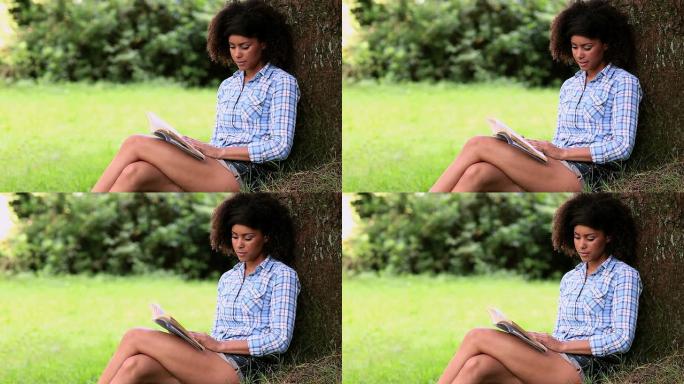 美丽的女人靠在树上在公园里读书