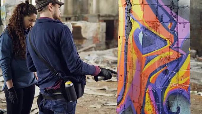 城市艺术家英俊的大胡子男人正在教业余学生使用气溶胶涂料，同时用抽象涂鸦装饰旧工业建筑。