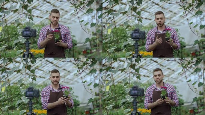 年轻微笑的博主男子花店在围裙拿着花说话，并为他的在线园艺视频博客录制视频博客