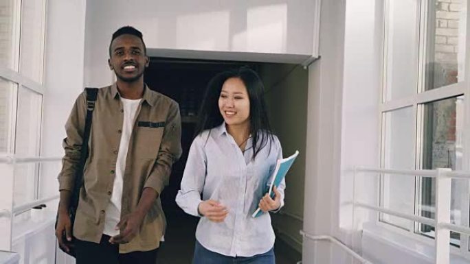 年轻英俊的非洲裔男学生和他的女朋友正走在大学走廊上讨论一些事情。他们遇到朋友站在窗户附近挥舞着他们