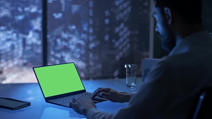 深夜，商人在他的私人办公室里用绿色模拟屏幕的笔记本电脑工作，可以看到大城市的窗户。