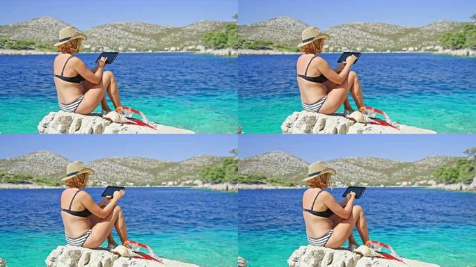 穿着比基尼的女人在阳光明媚的夏日蓝色海洋上使用数字平板电脑在岩石上