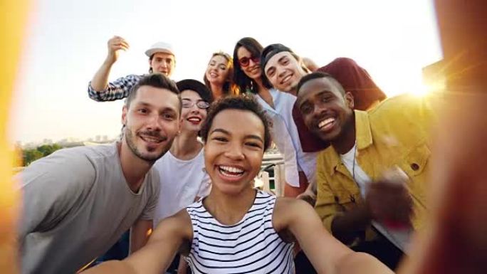 非裔美国女孩拿着相机，在屋顶派对上与快乐的朋友自拍的视点镜头。男人和女人看着相机，摆姿势大笑。