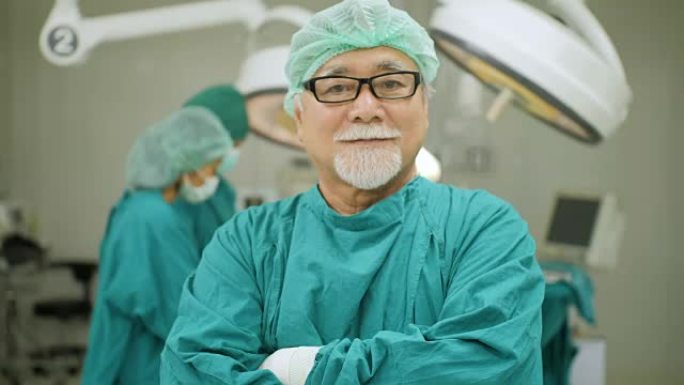 高级男性外科医生的肖像戴着全手术磨砂膏微笑的相机，团队医生在医院手术室为病人做手术。