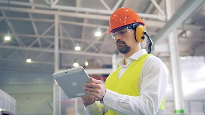 一位戴着安全帽和头戴式耳机的工人在他的小工具上键入信息，在仓库中查看货物。