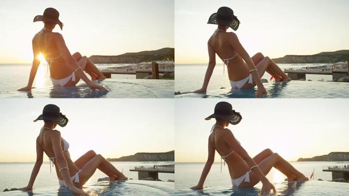 特写: 女人坐在无边泳池的边缘，在日落时看着海洋