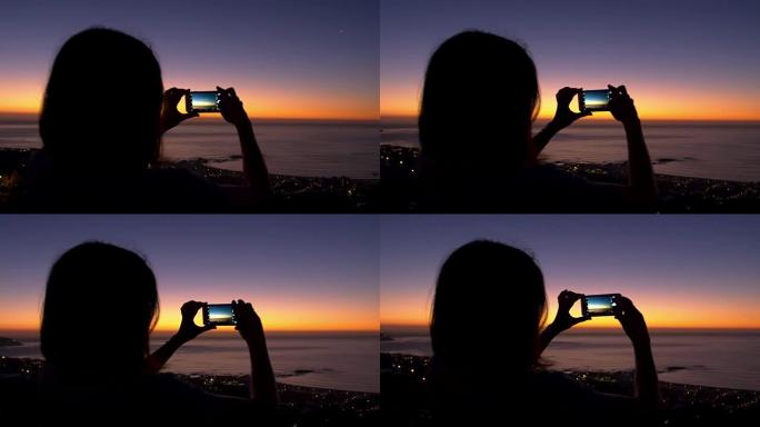 女人在海滩上用手机拍照，日落时剪影