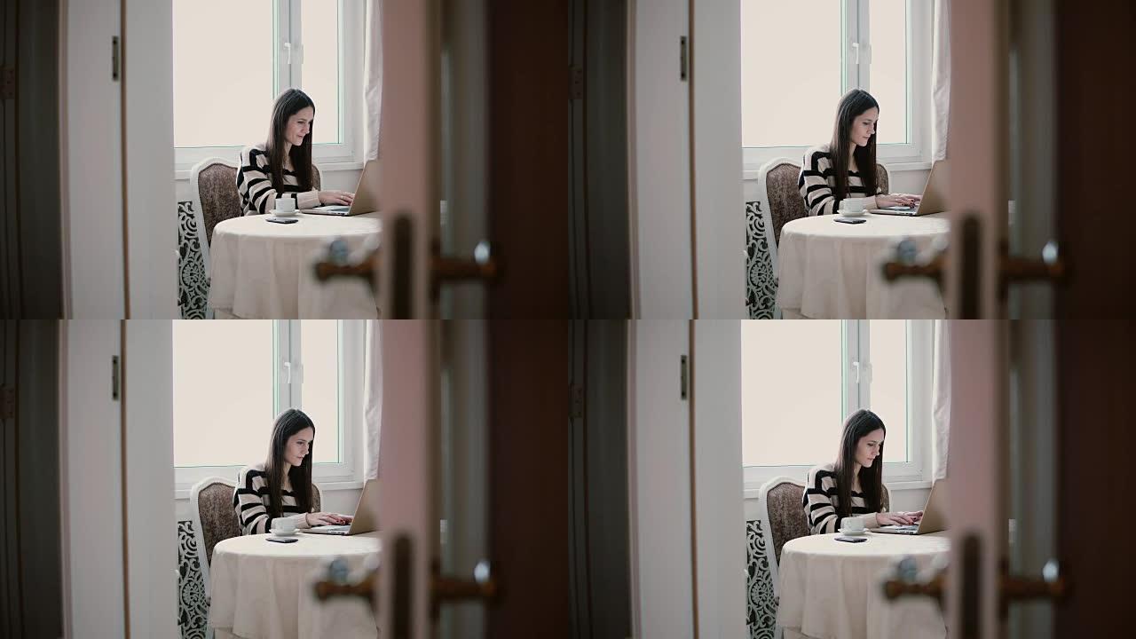 女人使用笔记本电脑，在明亮的用餐中享受早晨的咖啡。透过敞开的门观看