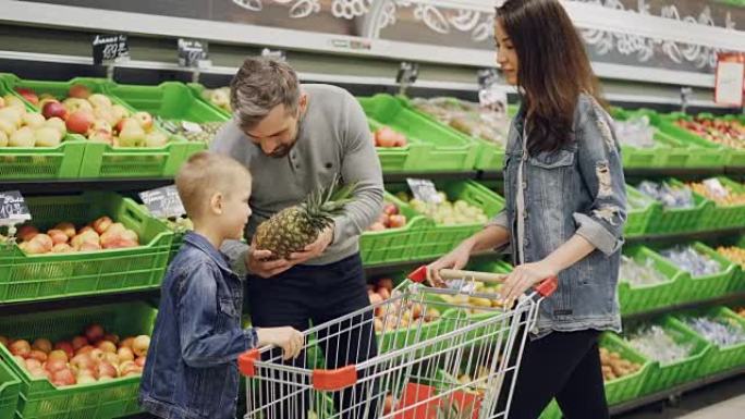 父亲，母亲和孩子在杂货店里选择菠萝，抚摸着它，闻到它的赞许。可以看到带有美味水果和购物手推车的架子。