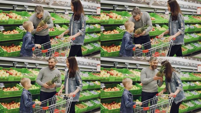 父亲，母亲和孩子在杂货店里选择菠萝，抚摸着它，闻到它的赞许。可以看到带有美味水果和购物手推车的架子。