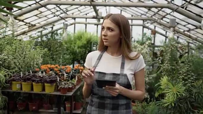 多莉拍摄了在花园中心工作的年轻女子。迷人的女孩在温室工作期间使用平板电脑检查和计数花朵