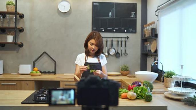 年轻的亚洲女子在厨房里用相机录制视频。微笑的亚洲女性致力于厨房里的水果和蔬菜的食物博客概念。