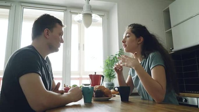 年轻的高加索夫妇坐在现代宽敞的厨房里的餐桌旁，以积极的方式讨论一些东西，他们正在喝茶，微笑，女人很漂