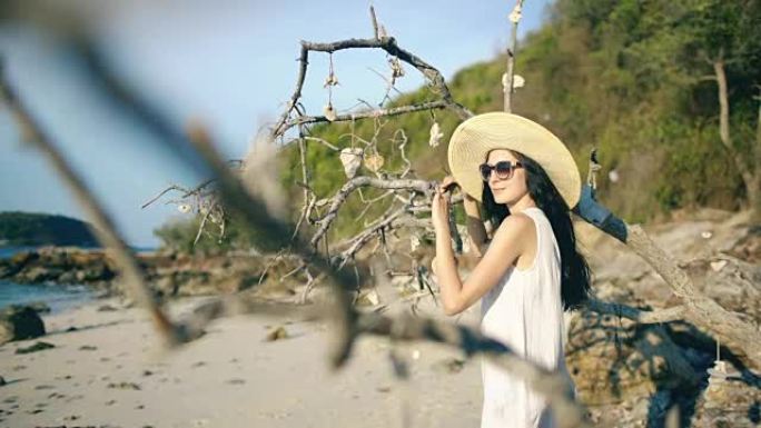 年轻性感的女人戴着太阳镜和帽子微笑着，在美丽的热带海滩的树附近摆姿势
