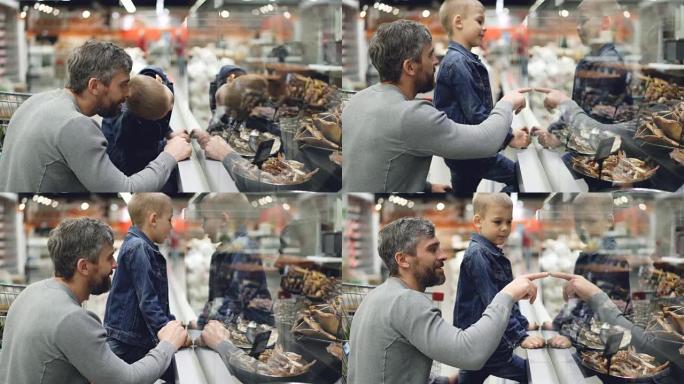 父亲和小儿子在超市里透过玻璃看食物，男人指着产品，笑着和儿子说话，好奇的男孩在微笑。