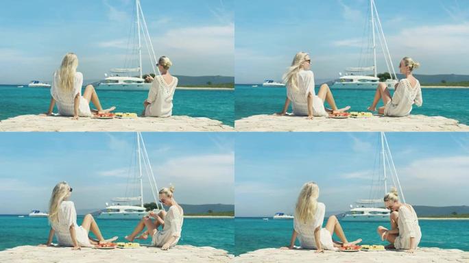 海边两个美丽的金发女孩坐在码头上，从托盘上吃水果，看着帆船经过。Torquise和天空的海洋颜色是清