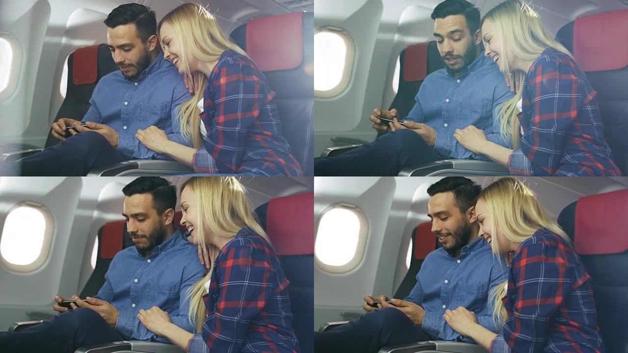 在商用飞机上，漂亮的年轻金发女郎与英俊的西班牙裔男性在智能手机上观看视频并微笑。阳光透过飞机的窗户照