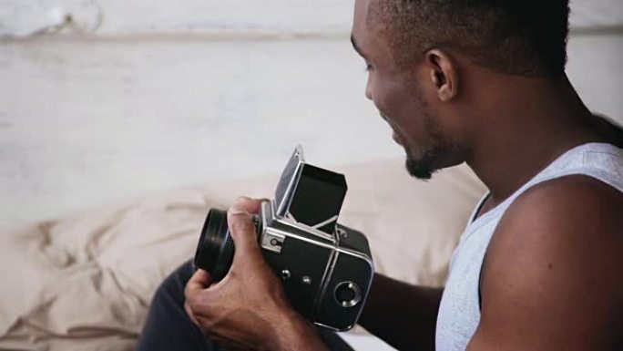 穿着睡衣的非洲年轻人坐在床上，拿着旧的照相机。移动胶片相机上的家伙说话照片