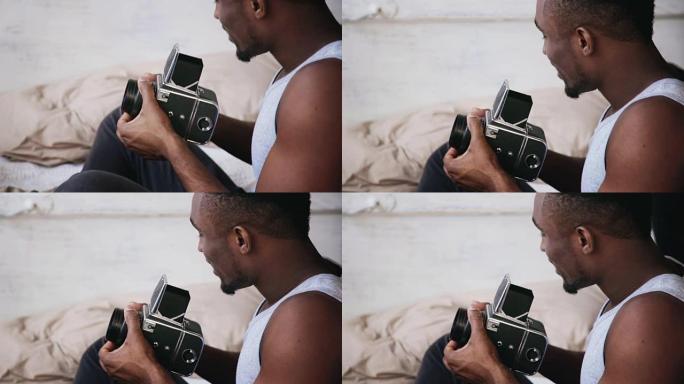 穿着睡衣的非洲年轻人坐在床上，拿着旧的照相机。移动胶片相机上的家伙说话照片