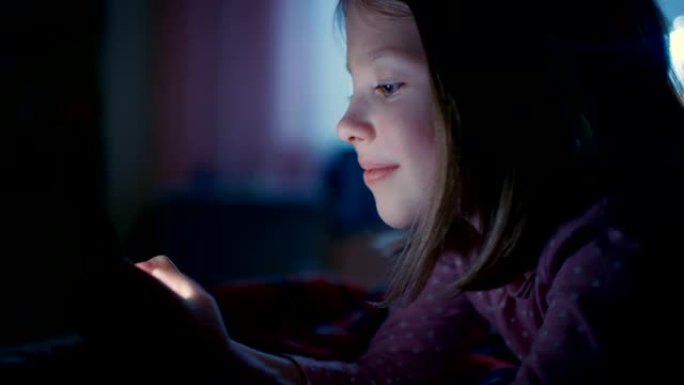 可爱的小女孩晚上在她的房间里，躺在床上使用智能手机。屏幕照亮了她的脸。
