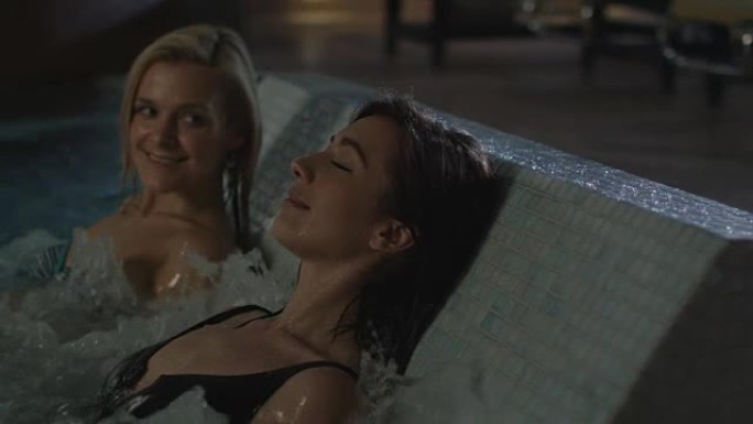 两个迷人的女孩正在水疗健康中心的热水浴缸上的游泳池里放松。