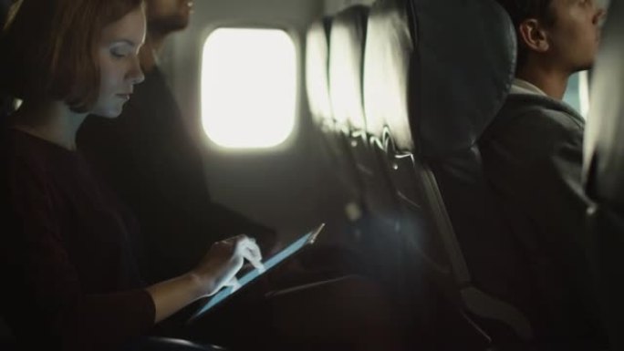 年轻女子正在飞机上使用平板电脑，一名男子正在窗户旁边的背景中听音乐。