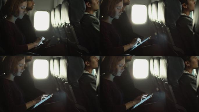 年轻女子正在飞机上使用平板电脑，一名男子正在窗户旁边的背景中听音乐。