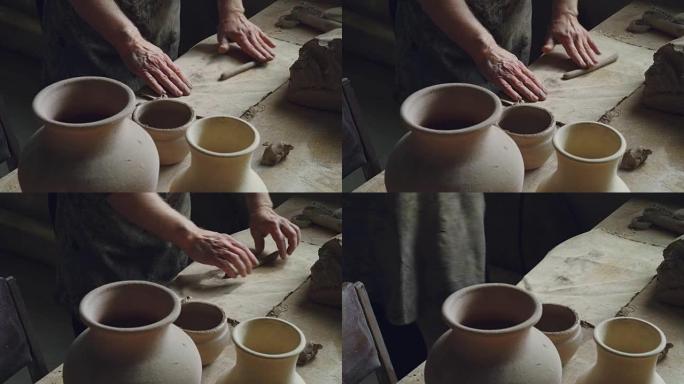 高级陶艺家的脏手正在工作台上滚动一小块粘土。手工制作的陶罐和罐子，波特的工具是可见的。