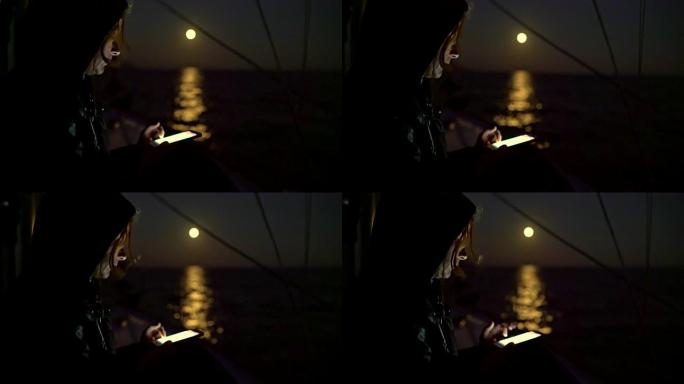 4k女人晚上在帆船上使用智能手机，满月在背景中照耀着海洋，实时