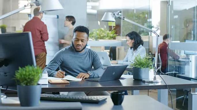 繁忙的国际办公室，非裔美国人在笔记本电脑上的办公桌前工作，他提醒自己的笔记本。背景富有创造力的年轻人