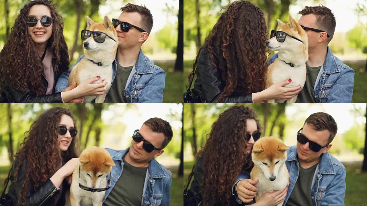 骄傲的主人给可爱的柴犬小狗戴上墨镜，夏天在城市公园里笑着聊天放松。人、动物、自然和有趣的概念。
