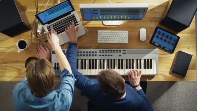 两位音频工程师在阳光工作室工作的俯视图。他们在音乐键盘上演奏并尝试声音。