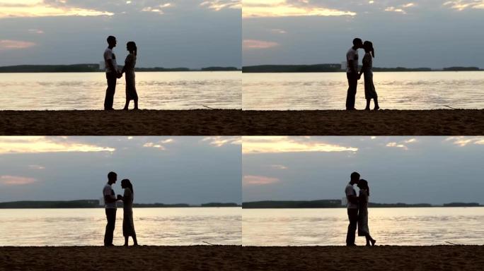 日落时一对恋爱中的年轻夫妇的剪影。女孩踮起脚尖，轻轻地亲吻湖岸上的那个家伙