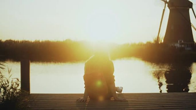女人坐在一个小湖码头上，看着日落。黄金时段。和平与内心和谐的完美时刻。4k后视图
