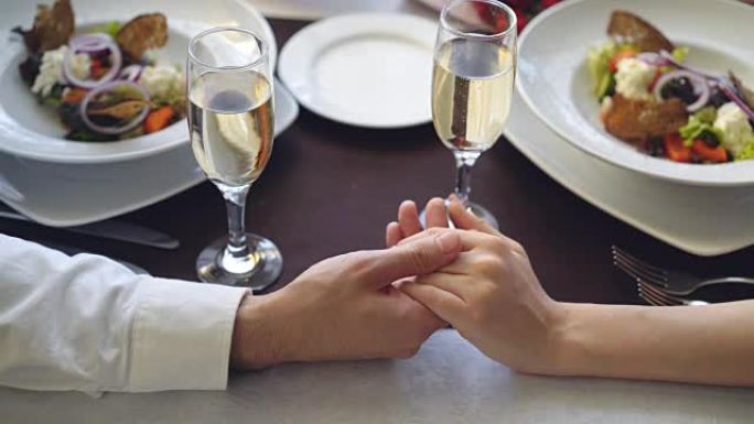 用香槟杯和盘子在桌子上握住和爱抚女性手的特写镜头。浪漫的关系，爱情和精致的餐饮概念。