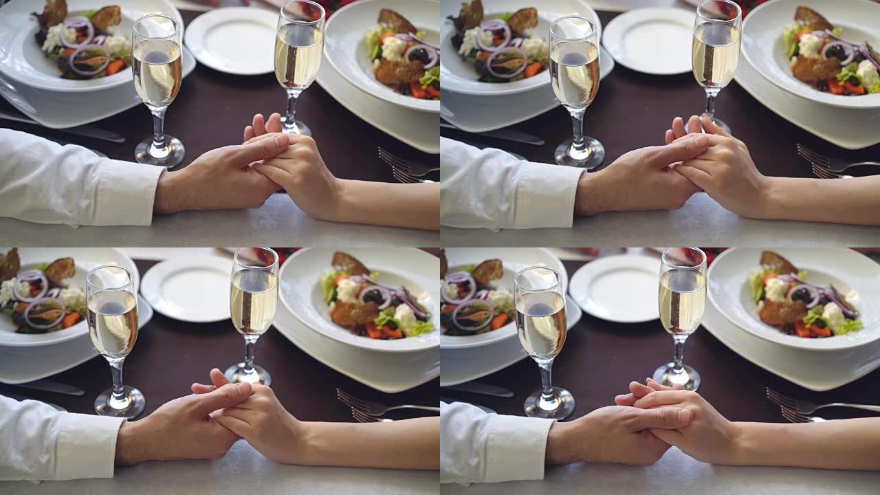 用香槟杯和盘子在桌子上握住和爱抚女性手的特写镜头。浪漫的关系，爱情和精致的餐饮概念。