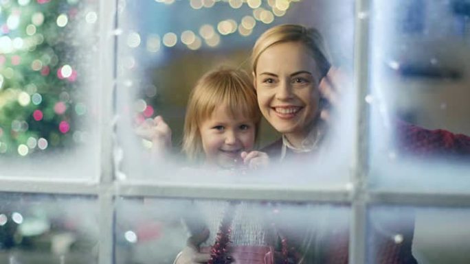 在圣诞节前夕，母女俩透过白雪皑皑的窗户，对着镜头挥手致意。圣诞Lites在窗户上闪闪发光。