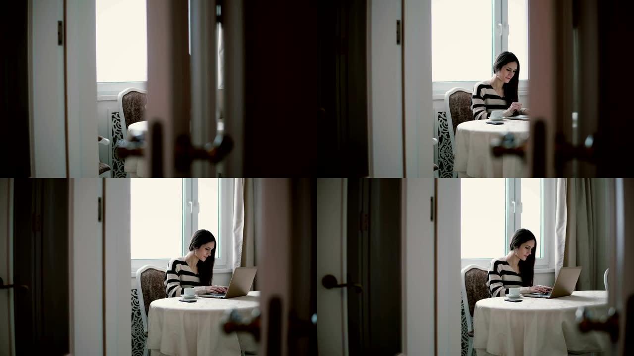 女人使用笔记本电脑，在明亮的用餐中享受早晨的咖啡。向左滑动，通过打开的门查看