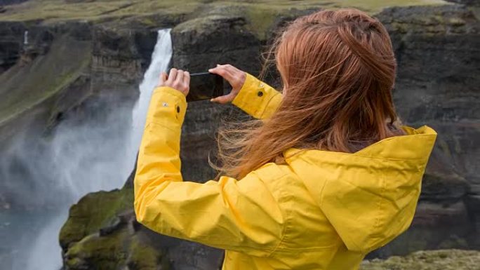 女人站在悬崖上，用智能手机拍摄瀑布的照片