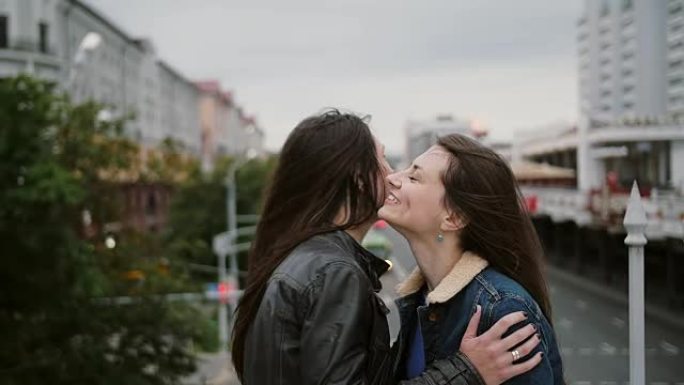 两个漂亮的女孩在城市桥上相遇，拥抱，交谈，微笑，大笑。风吹他们长发