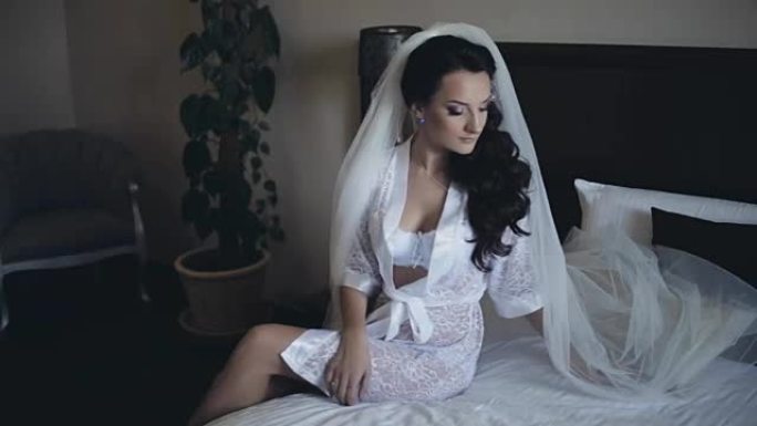 年轻迷人的新娘穿着优雅的白色内衣，peignoir坐在床上。黑发女人固定她的面纱和姿势