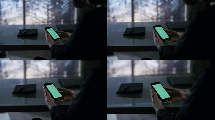 女商人坐在办公桌前，使用带有绿屏的智能手机。窗外可以看到大城市。