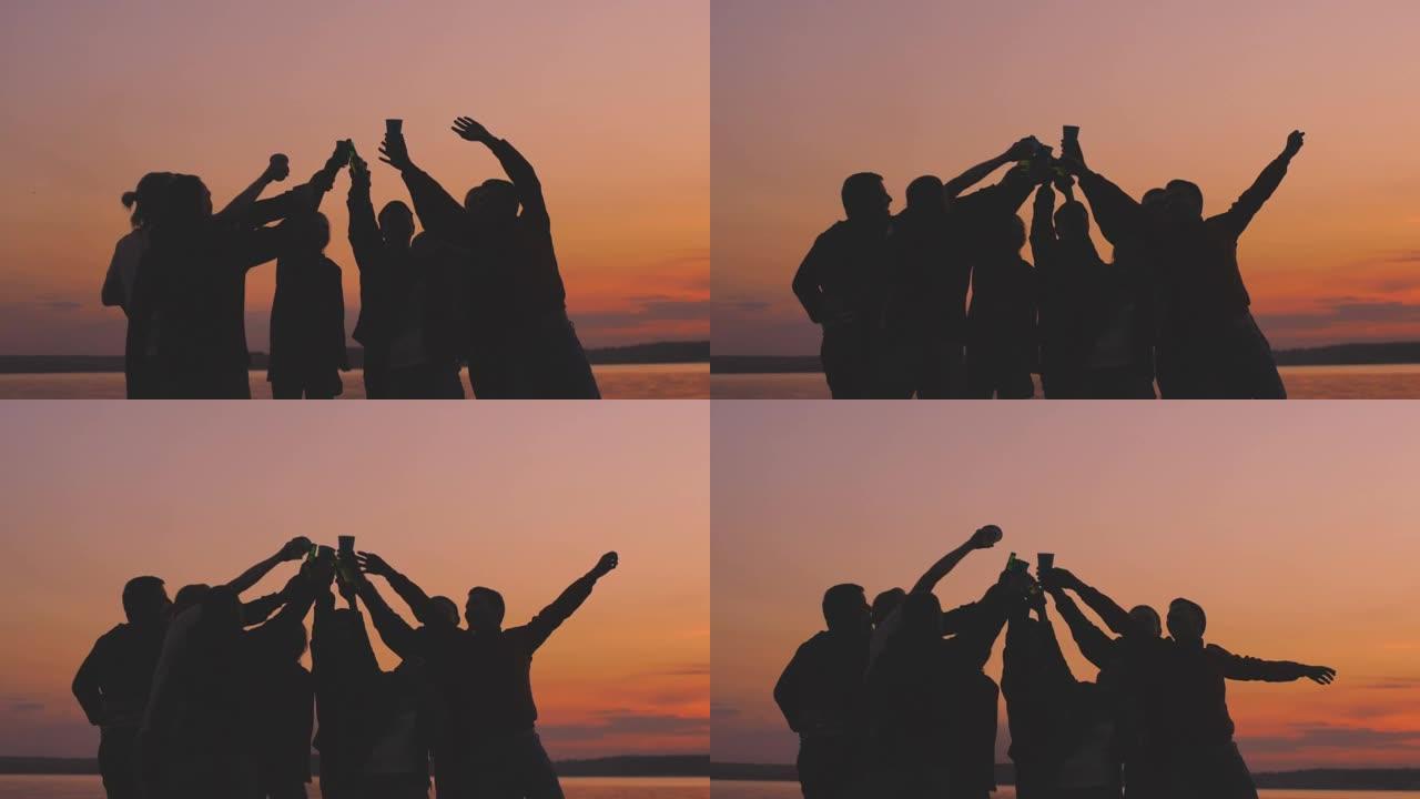 剪影组的慢动作年轻跳舞的人们在日落时在海滩上欢呼并举行聚会