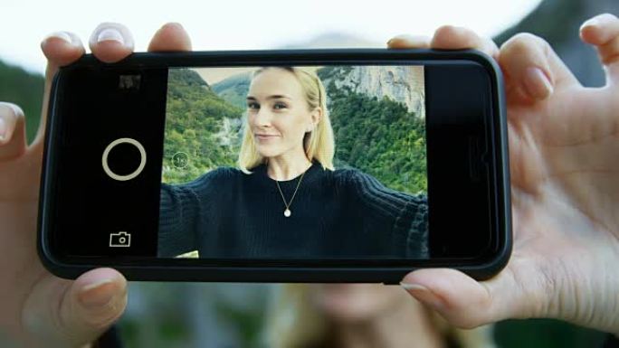 智能手机屏幕上美丽的金发女孩自拍的特写镜头。在背景风景秀丽的青山山景。