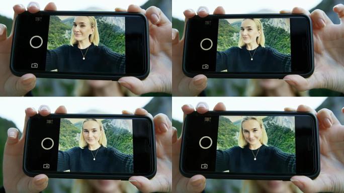 智能手机屏幕上美丽的金发女孩自拍的特写镜头。在背景风景秀丽的青山山景。