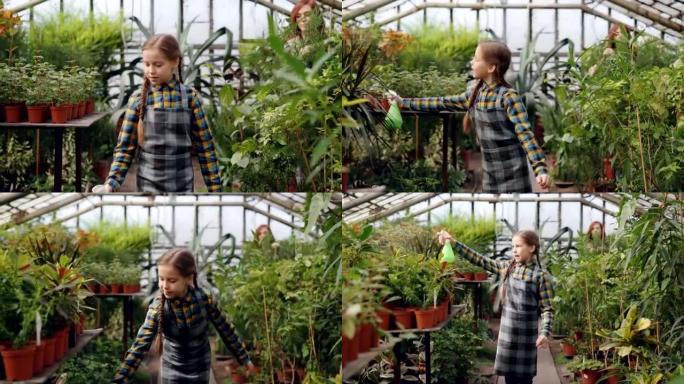 漂亮女孩在温室里散步，在绿色植物上喷水，而她忙碌的母亲在后台工作。童年，帮助父母和家庭观念。
