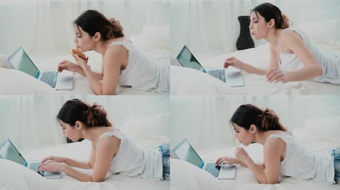 美丽的女孩早上躺在床上，用笔记本电脑。少妇吃羊角面包打字上网
