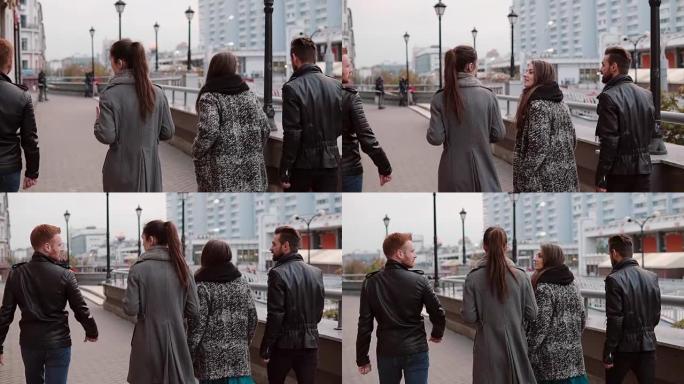 两个时髦的女孩和两个时髦的男人兴高采烈地走在城市里聊天。慢镜头，稳定镜头，后视图