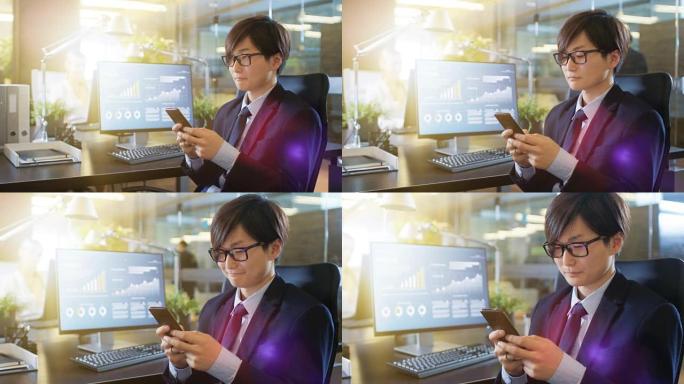 在办公室里，东亚商人使用智能手机，键入重要信息。他的台式计算机显示了公司发展的数据。阳光普照。