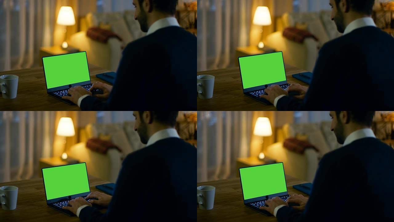 在家里，一个男人坐在办公桌前，在笔记本电脑上打字，上面放着绿屏。他的公寓是黄色的，很温暖。